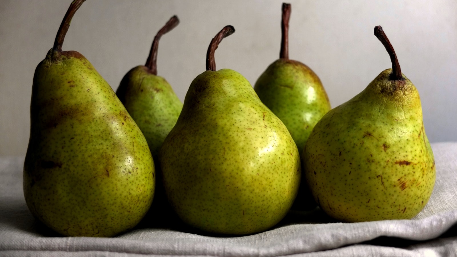 Five green pears arranged in a row on a pale linen cloth.