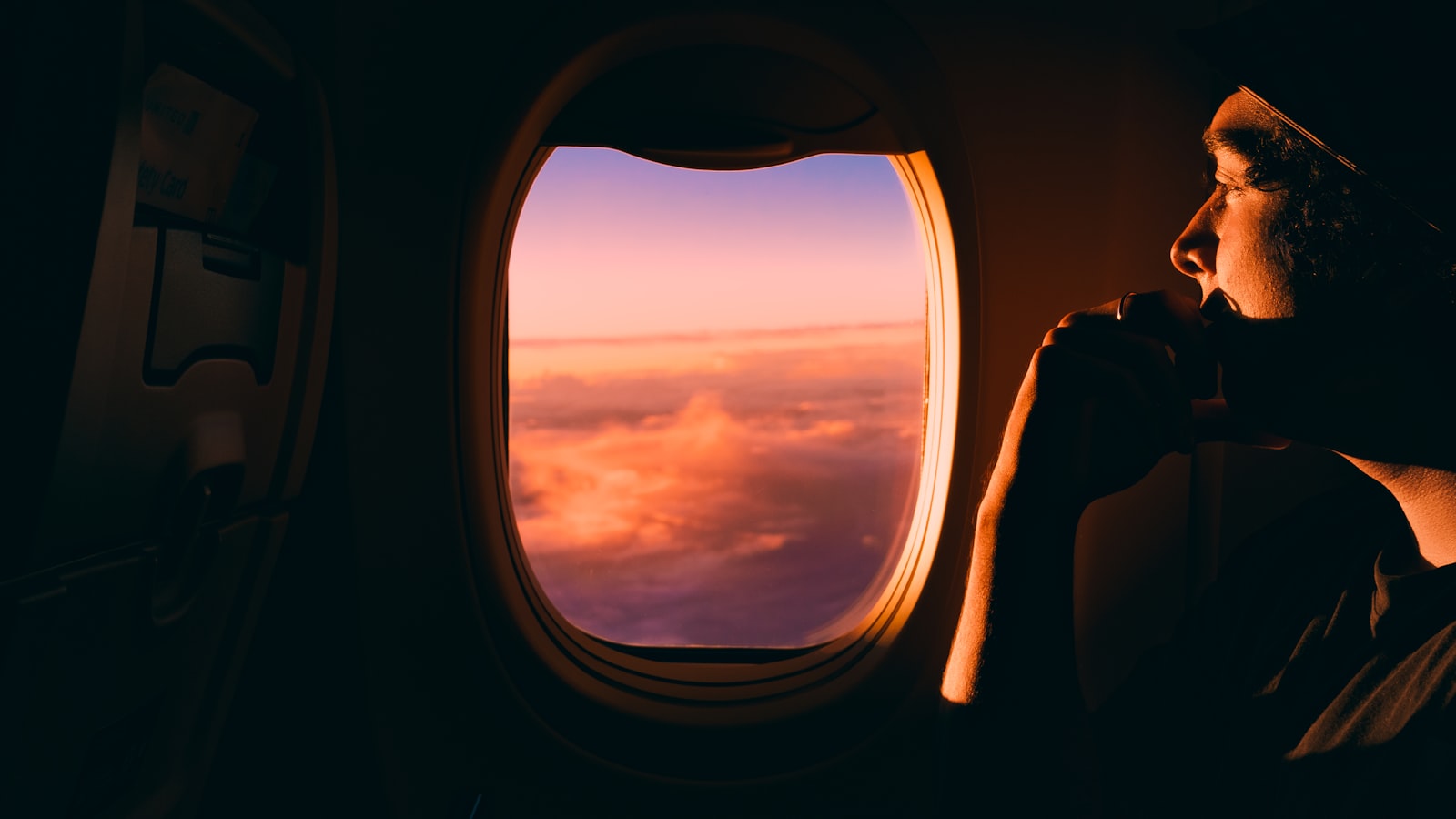 A young person gazes quietly out of an airplane window at warm sunset light.