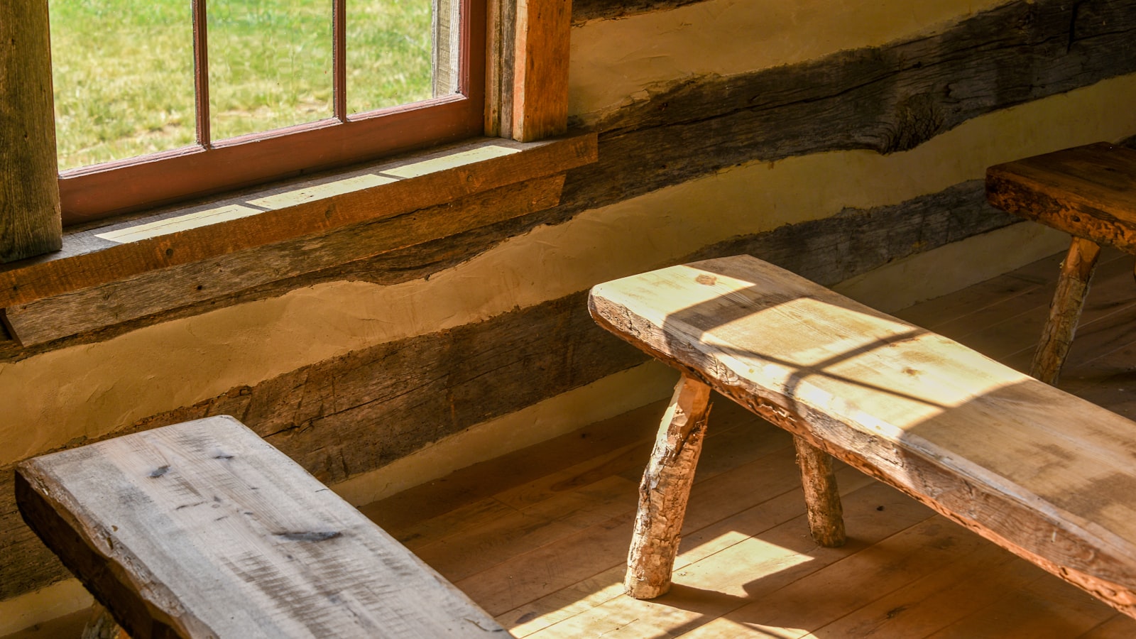 A worn wooden bench sits by a quiet window in a rustic cabin, sunlight pouring through the panes.