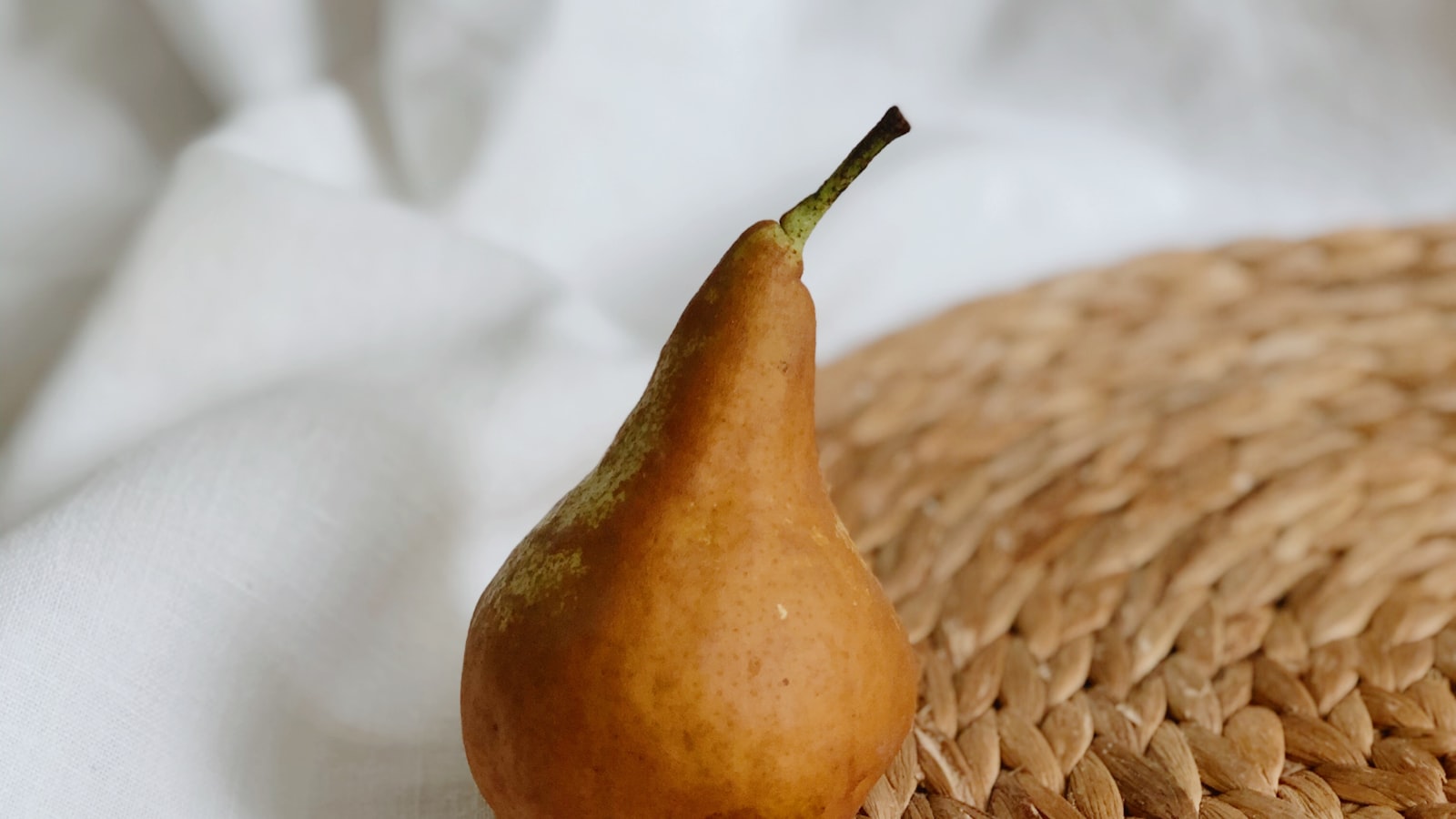 A single ripe pear sitting on a woven mat beside softly draped linen.