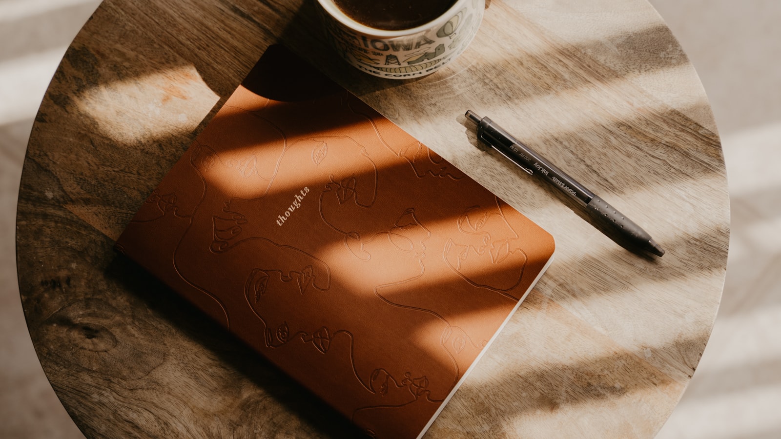 A warm terracotta notebook on a round wooden table, sunlight from window blinds striping across it.